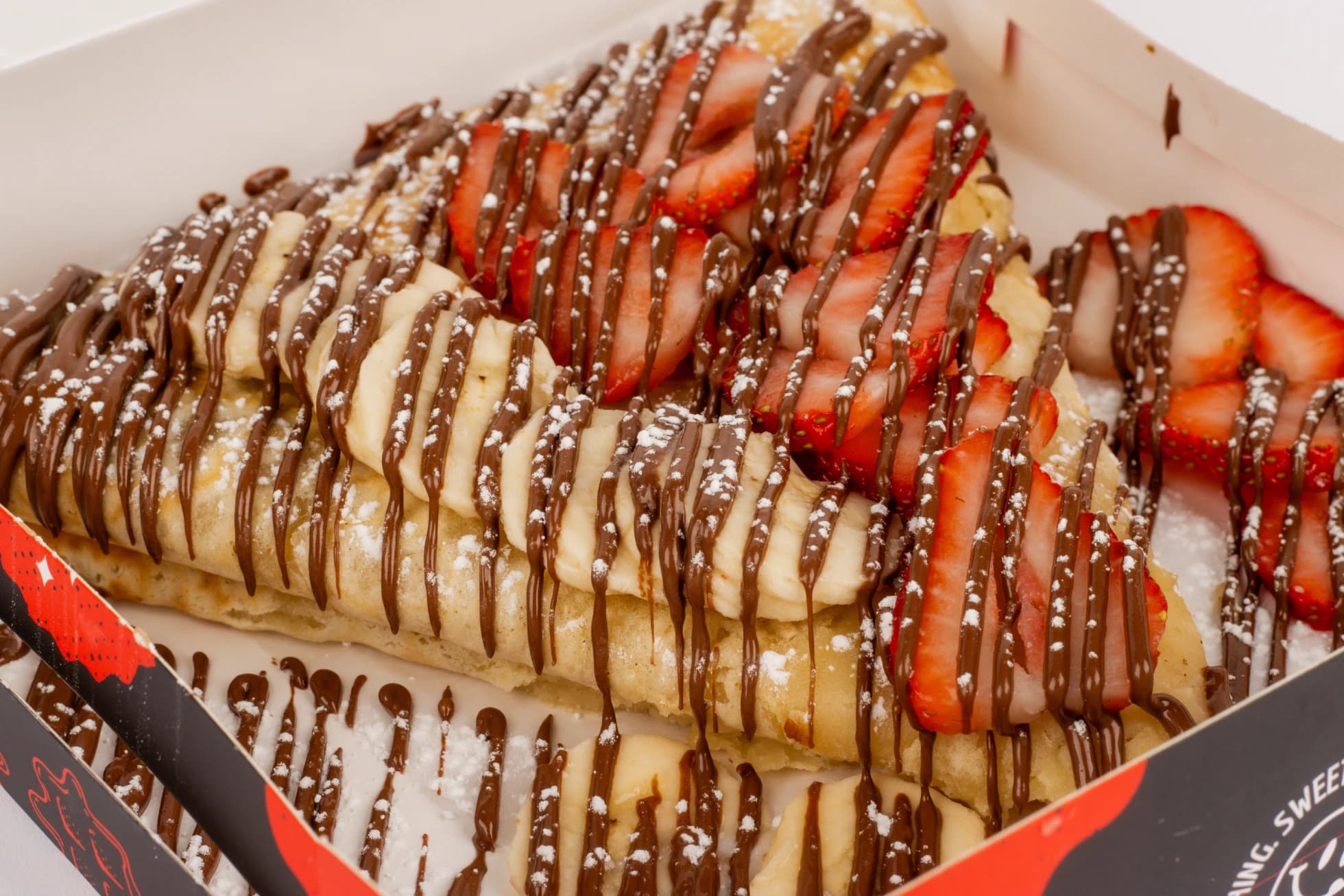 A Battr catering box of flat crepes layered with sliced strawberries and bananas, drizzled with Nutella and dusted with powdered sugar.