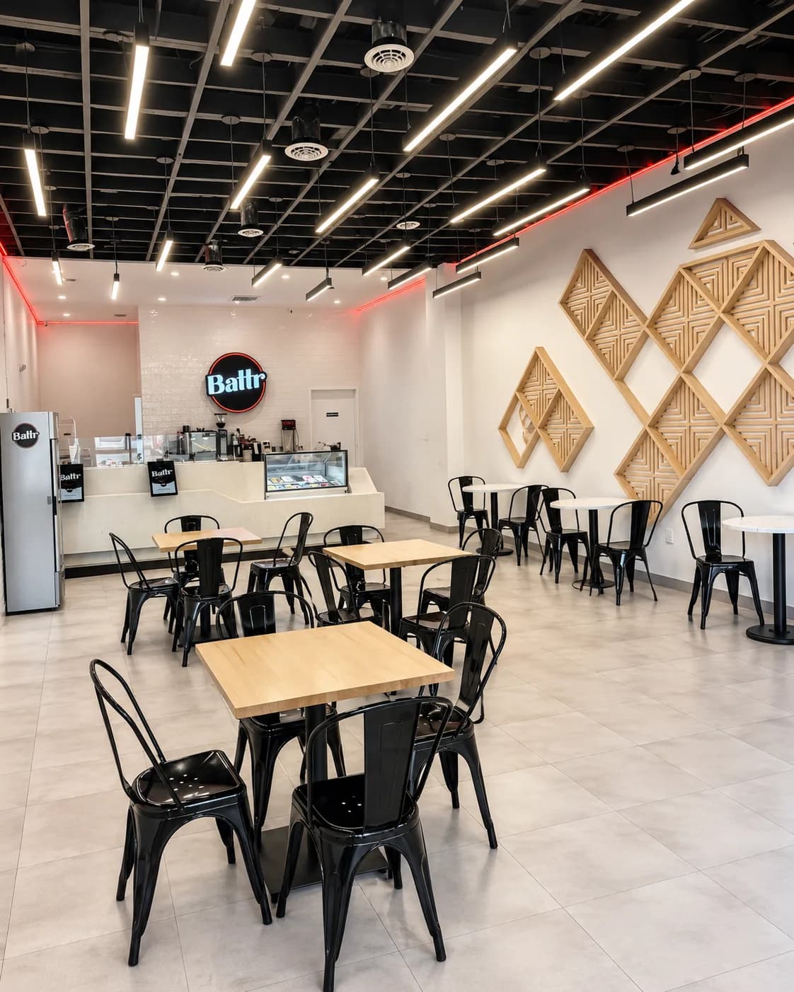 The dining room at Battr Lakewood — black bistro tables and chairs on pale tile, a lit Battr sign above the white-tiled counter, and a wall of diamond-shaped wooden panels.