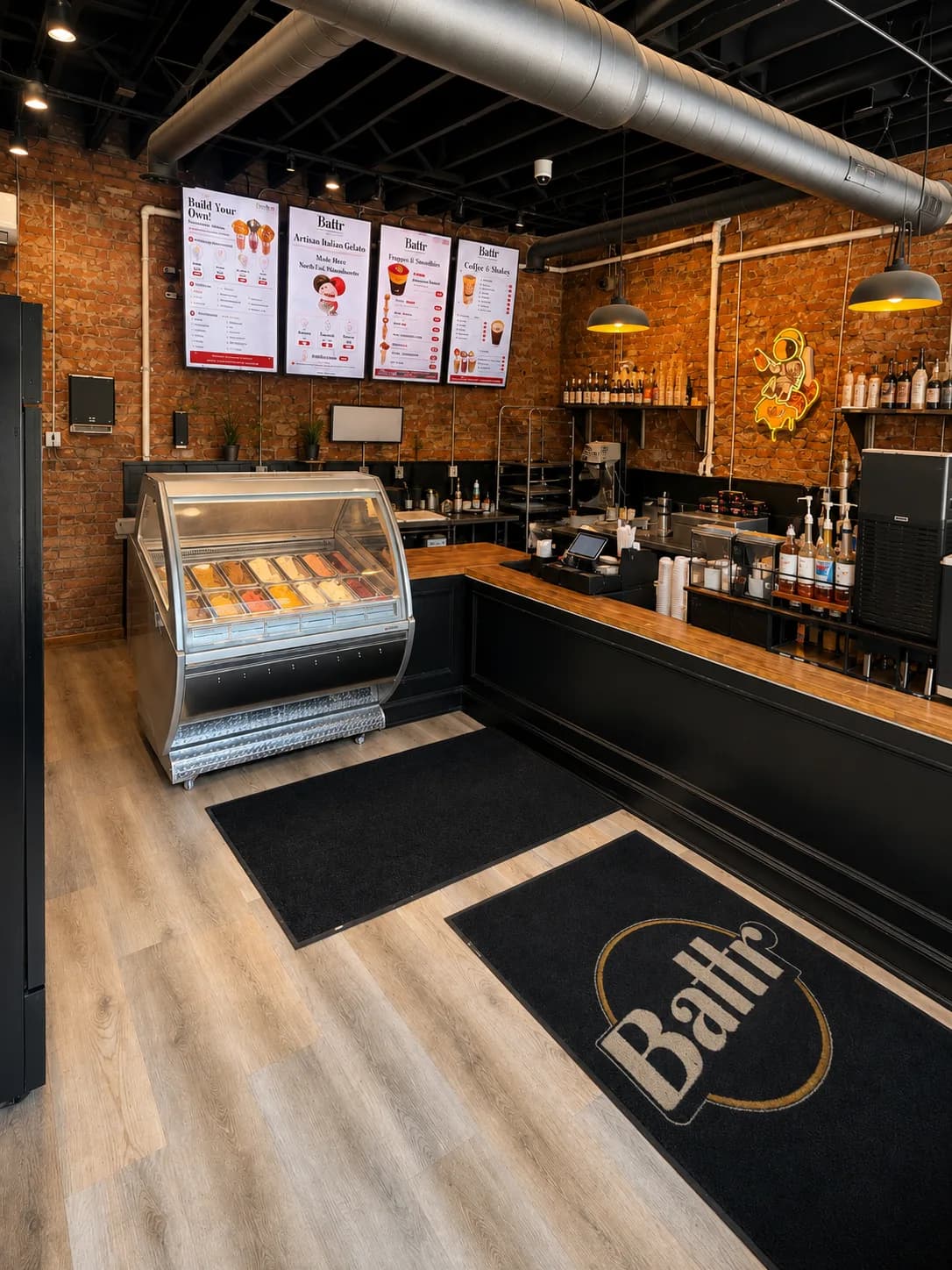 The counter at Battr Tremont — menu boards on the exposed brick wall, a curved gelato display case, the espresso bar with syrup bottles, and a branded Battr floor mat on the wood floor.